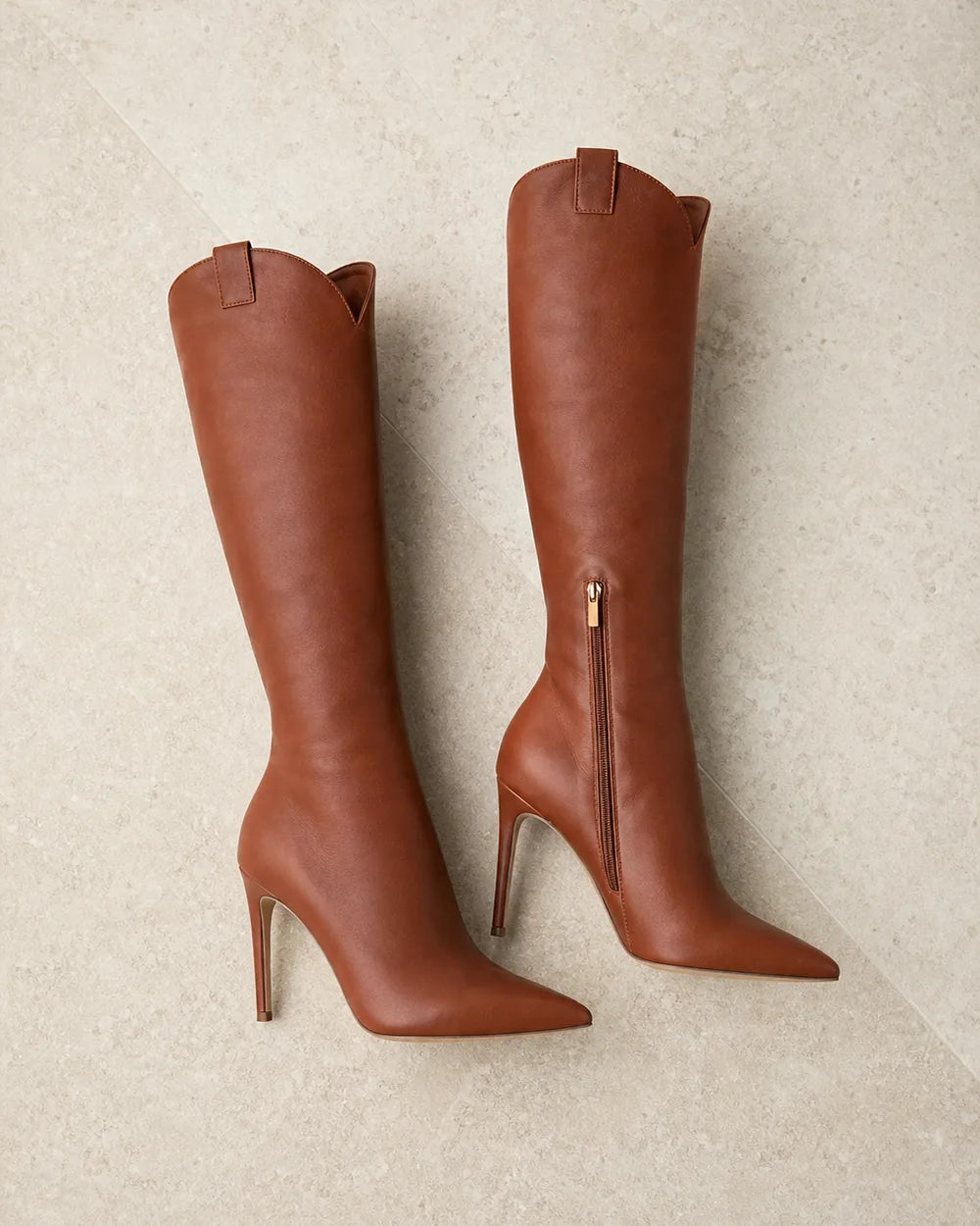 Women's brown leather knee-high stiletto boots arranged on pale stone.